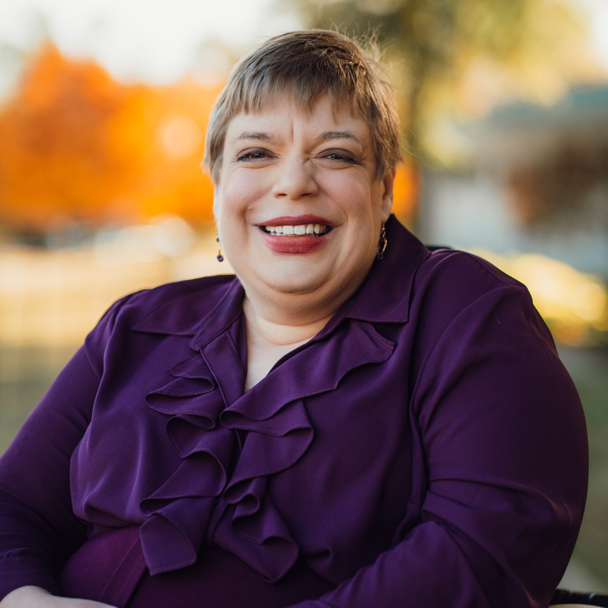 A professional woman with short hair and a warm smile sits outdoors, wearing a deep purple blouse with ruffled detail. She is seated comfortably with her hands folded, and soft autumn foliage and trees appear blurred in the background, creating a warm, natural setting.